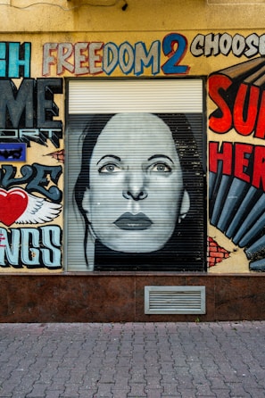 A wall features a mural with vibrant graffiti art. At the center is a monochrome portrait of a woman's face on a roller shutter. Surrounding the portrait are colorful words and designs, including the word 'freedom' painted in bold, diverse colors. The artwork juxtaposes words and images, creating a dynamic urban landscape.