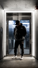 A person dressed in dark clothing and a cap stands in front of an illuminated elevator entrance. The elevator background is metallic with a sign displaying the hours of operation. The scene is dimly lit with a modern, urban feel.