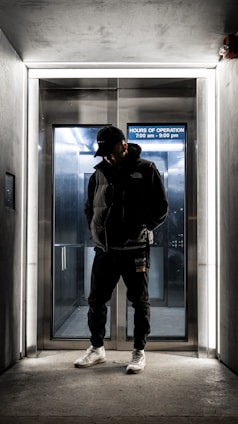 A person dressed in dark clothing and a cap stands in front of an illuminated elevator entrance. The elevator background is metallic with a sign displaying the hours of operation. The scene is dimly lit with a modern, urban feel.