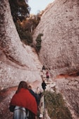 A group of people is hiking up a narrow stone pathway surrounded by large, rocky cliffs. The path is steep and bordered by a railing. Dense greenery and trees are visible at the top of the cliffs, indicating a natural and rugged terrain.