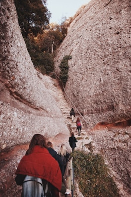 A group of people is hiking up a narrow stone pathway surrounded by large, rocky cliffs. The path is steep and bordered by a railing. Dense greenery and trees are visible at the top of the cliffs, indicating a natural and rugged terrain.