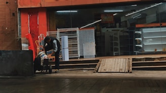 A person is leaning over a cart in front of a store with a glass facade. The store appears closed and has empty shelves visible inside. Several metal racks and carts are positioned outside against a red painted wall.