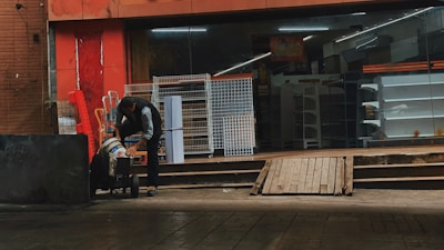 A person is leaning over a cart in front of a store with a glass facade. The store appears closed and has empty shelves visible inside. Several metal racks and carts are positioned outside against a red painted wall.