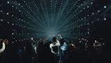 A crowd of people, many holding up smartphones, watching a light show with beams radiating from the center of a dark room. The arrangement of lights creates a tunnel effect, drawing focus towards the distant center point.