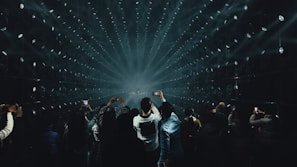 A crowd of people, many holding up smartphones, watching a light show with beams radiating from the center of a dark room. The arrangement of lights creates a tunnel effect, drawing focus towards the distant center point.
