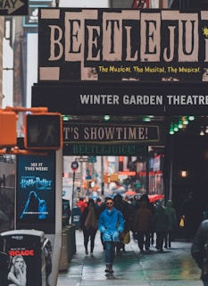 A busy city street scene with pedestrians walking towards the camera. Above them is a prominent theater marquee advertising 'Beetlejuice The Musical' at the Winter Garden Theatre. The atmosphere is lively with signs and lights all around, indicating a bustling urban environment.