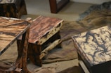 Wooden stools with intricate, multi-colored patterns on their surfaces. The stools have a natural, rustic look and are placed on worn fabric or bedding material.