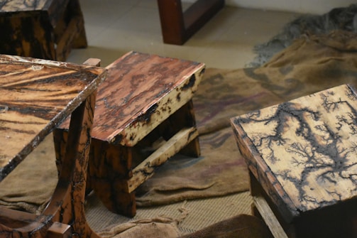 Wooden stools with intricate, multi-colored patterns on their surfaces. The stools have a natural, rustic look and are placed on worn fabric or bedding material.