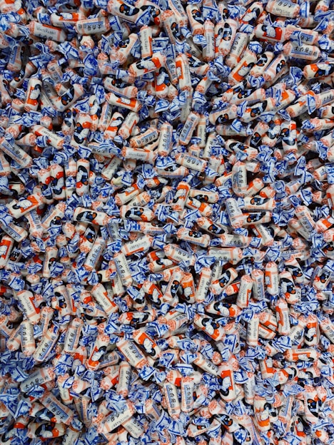 A large pile of colorful candies wrapped in white, blue, and orange packaging. The candies have illustrations and text on them, showcasing a playful and vibrant design.