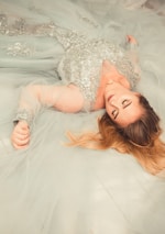 A woman lies on a soft, flowing, light blue gown with intricate silver embellishments. Her eyes are closed, and her long blonde hair is spread out around her.