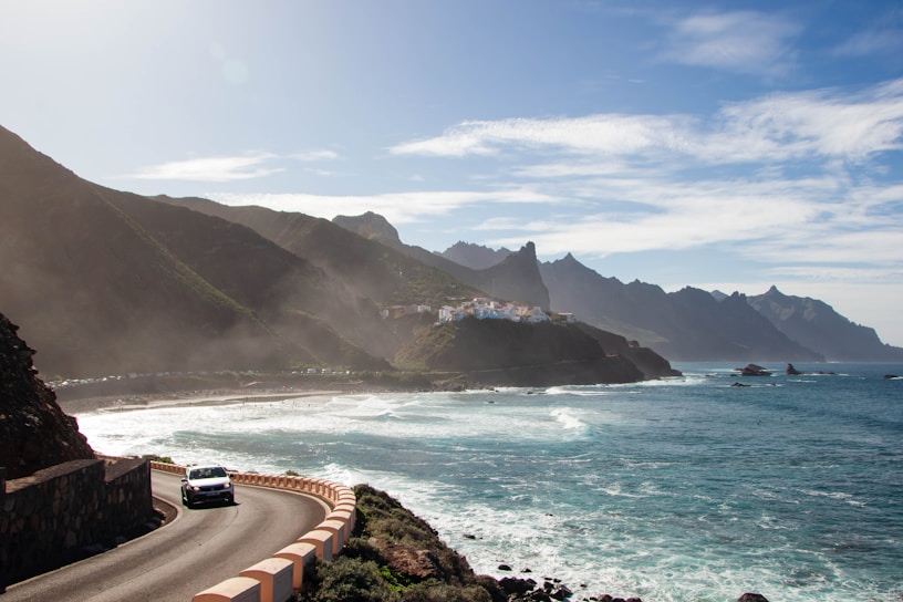 A scenic coastal landscape featuring a winding road along a cliffside with a car traveling on it. To the left, towering mountains create a dramatic backdrop with misty air near the peaks. To the right, the ocean extends with foamy waves crashing against the rocks. In the middle distance, a small village with white buildings is nestled against the hills.