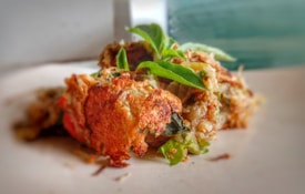 A close-up image of a savory dish featuring a crispy, golden brown exterior with visible herbs and vegetables. The dish is garnished with fresh green leaves, and the texture looks flaky and appetizing.