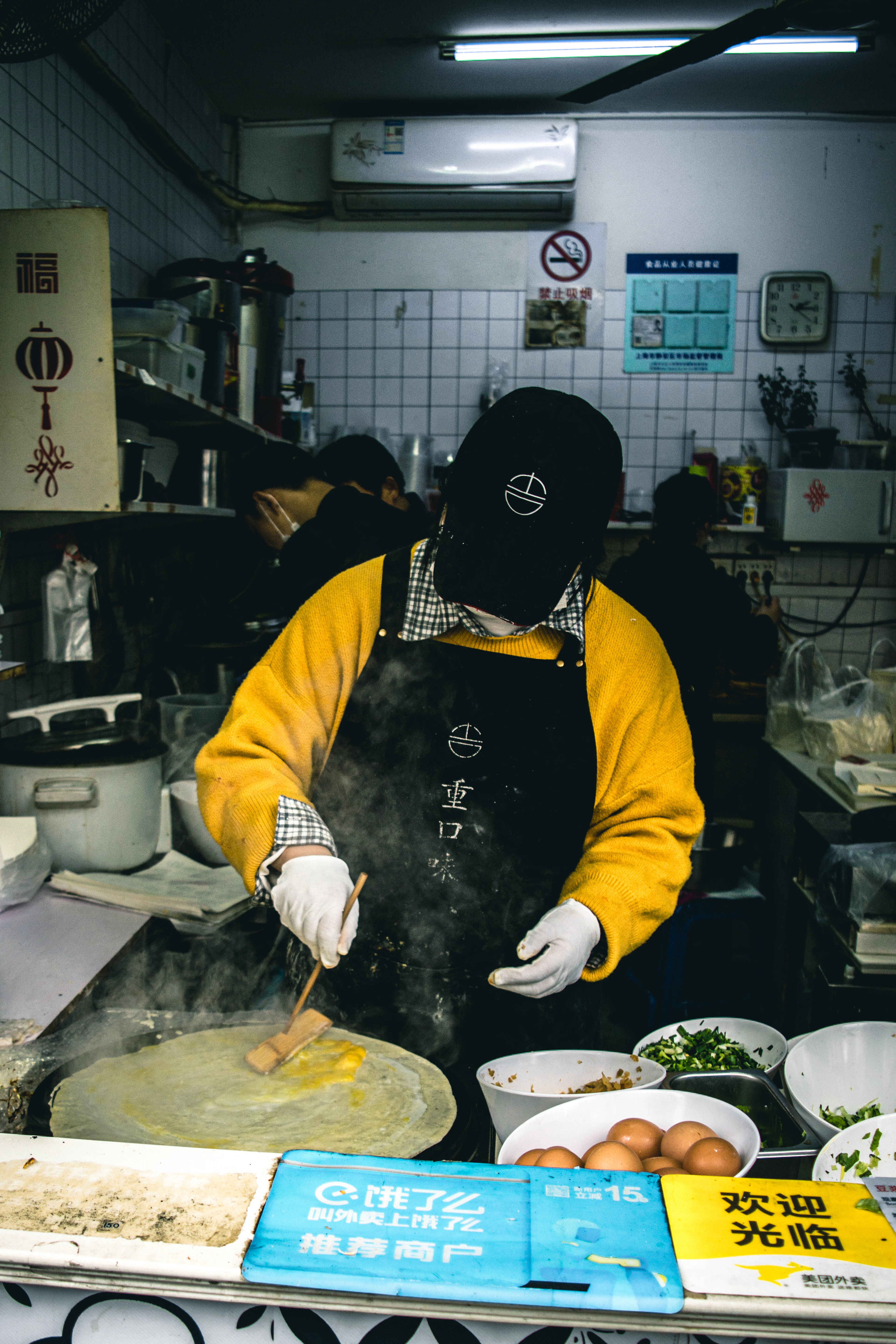 A street cook spreading batter for a jianbing crepe across a hot griddle, with bowls of chopped scallion, eggs and sauces at his side, at a Shanghai street-food stall