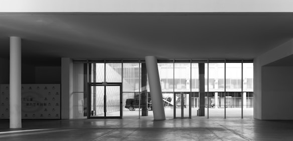 An architectural space featuring a modern entrance with multiple glass doors and tall windows. The area includes distinct angled columns and an exterior view showing a parked vehicle and building structures in a minimalist style. The scene is primarily monochromatic, emphasizing structural lines and simplicity.