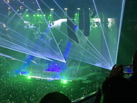 A concert scene with a crowd of people at the bottom, a stage in the center featuring musicians, and vibrant lights and laser effects in green and blue. The environment is dark, with bright laser beams crisscrossing across the stage area. Two large screens or panels are visible behind the stage, enhancing the visual effects.