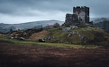 A medieval stone castle ruin sits atop a grassy hill, surrounded by a rugged landscape with rolling hills in the background. Dark gray clouds loom overhead, creating a dramatic and gloomy atmosphere. Several off-road vehicles are parked on the grass nearby, adding a modern element to the historical setting.