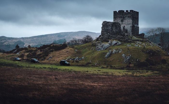 A medieval stone castle ruin sits atop a grassy hill, surrounded by a rugged landscape with rolling hills in the background. Dark gray clouds loom overhead, creating a dramatic and gloomy atmosphere. Several off-road vehicles are parked on the grass nearby, adding a modern element to the historical setting.