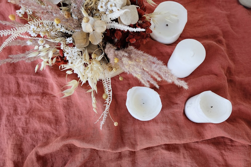 A collection of dried flowers and foliage are artistically arranged atop a textured, warm-toned fabric. Surrounding the arrangement are several white candles, some of which show signs of being previously lit. The combination of natural elements and soft candle wax creates a serene and rustic atmosphere.