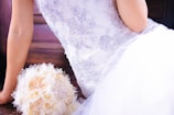 An elegantly dressed person in a lace-embellished white gown is sitting beside a bouquet made from fabric and lace, featuring pastel tones. The gown has intricate floral patterns, and the scene conveys a sense of bridal beauty.