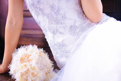 An elegantly dressed person in a lace-embellished white gown is sitting beside a bouquet made from fabric and lace, featuring pastel tones. The gown has intricate floral patterns, and the scene conveys a sense of bridal beauty.
