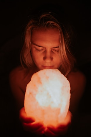 A person with closed eyes holds a glowing, orange-hued object that appears to be a salt lamp close to their face. The background is dark, emphasizing the soft light emitted by the lamp and creating a warm, intimate atmosphere.