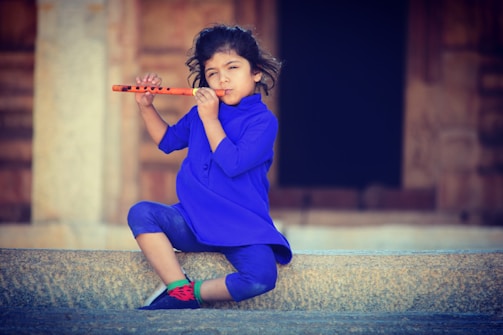 A young child is seated on stone steps, playing an orange flute. The child is dressed in a vibrant blue outfit with blue pants and a green sock visible alongside red shoes. The background shows a blurred wooden structure, possibly a door.