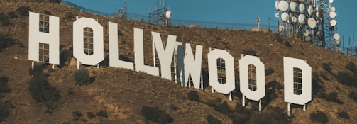 Large white letters spell out 'Hollywood' on a hillside covered in dry brown vegetation. Communication towers and antennas are visible in the background, set against a clear blue sky.
