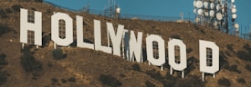 Large white letters spell out 'Hollywood' on a hillside covered in dry brown vegetation. Communication towers and antennas are visible in the background, set against a clear blue sky.