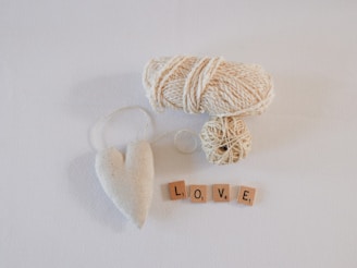 A spool of beige yarn, a beige heart-shaped ornament with a string, and four wooden tiles spelling out 'LOVE' are arranged on a white surface.