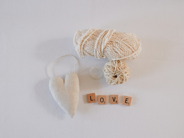 A spool of beige yarn, a beige heart-shaped ornament with a string, and four wooden tiles spelling out 'LOVE' are arranged on a white surface.