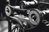An intricate assembly of gears and chains, highlighting the mechanical components of a machine. The photo is captured in black and white, emphasizing the metallic textures and details of the machinery.