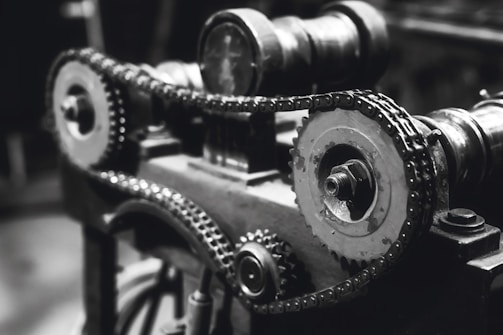 An intricate assembly of gears and chains, highlighting the mechanical components of a machine. The photo is captured in black and white, emphasizing the metallic textures and details of the machinery.