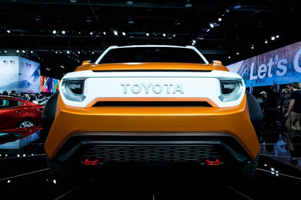 A front view of an orange Toyota car displayed at an auto show. The surroundings include bright lights, a crowd of people in the background, and various other vehicles and displays.