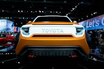 A front view of an orange Toyota car displayed at an auto show. The surroundings include bright lights, a crowd of people in the background, and various other vehicles and displays.