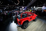 A vibrant red off-road vehicle with an open cabin layout is on display at a car show. The vehicle is parked on a wooden platform inside a large exhibition hall filled with people. Various car manufacturer logos and numerous other vehicles are visible in the background amid bright overhead lighting and display screens.