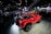A vibrant red off-road vehicle with an open cabin layout is on display at a car show. The vehicle is parked on a wooden platform inside a large exhibition hall filled with people. Various car manufacturer logos and numerous other vehicles are visible in the background amid bright overhead lighting and display screens.