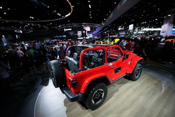 A vibrant red off-road vehicle with an open cabin layout is on display at a car show. The vehicle is parked on a wooden platform inside a large exhibition hall filled with people. Various car manufacturer logos and numerous other vehicles are visible in the background amid bright overhead lighting and display screens.