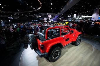 A vibrant red off-road vehicle with an open cabin layout is on display at a car show. The vehicle is parked on a wooden platform inside a large exhibition hall filled with people. Various car manufacturer logos and numerous other vehicles are visible in the background amid bright overhead lighting and display screens.