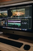 A computer monitor displays a video editing software interface with several video clips and sequences being edited. The workspace includes timelines, video previews, and various tools. Below the monitor are a keyboard and a mouse on a wooden desk.