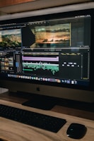A computer monitor displays a video editing software interface with several video clips and sequences being edited. The workspace includes timelines, video previews, and various tools. Below the monitor are a keyboard and a mouse on a wooden desk.
