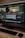 A computer monitor displays a video editing software interface with several video clips and sequences being edited. The workspace includes timelines, video previews, and various tools. Below the monitor are a keyboard and a mouse on a wooden desk.