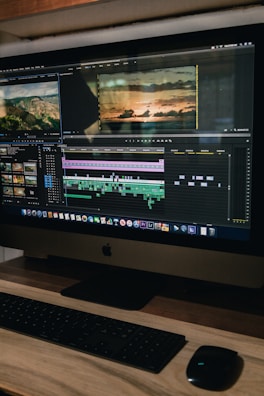 A computer monitor displays a video editing software interface with several video clips and sequences being edited. The workspace includes timelines, video previews, and various tools. Below the monitor are a keyboard and a mouse on a wooden desk.