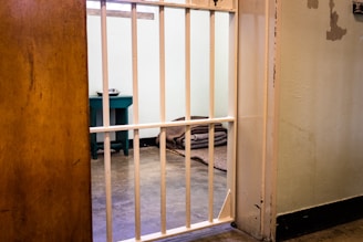 A small prison cell with metal bars partially open, revealing an interior that includes a narrow bed with a folded gray blanket and a small table holding a metal dish. The walls are bare, and the atmosphere is stark and unwelcoming.