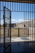 A prison cell door stands open, leading to an outdoor yard enclosed by high concrete walls. The sky is clear with a few clouds, and there is a metal fence surrounding the yard.