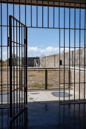 A prison cell door stands open, leading to an outdoor yard enclosed by high concrete walls. The sky is clear with a few clouds, and there is a metal fence surrounding the yard.