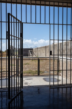 A prison cell door stands open, leading to an outdoor yard enclosed by high concrete walls. The sky is clear with a few clouds, and there is a metal fence surrounding the yard.