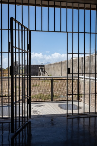 A prison cell door stands open, leading to an outdoor yard enclosed by high concrete walls. The sky is clear with a few clouds, and there is a metal fence surrounding the yard.