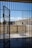 A prison cell door stands open, leading to an outdoor yard enclosed by high concrete walls. The sky is clear with a few clouds, and there is a metal fence surrounding the yard.