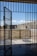 A prison cell door stands open, leading to an outdoor yard enclosed by high concrete walls. The sky is clear with a few clouds, and there is a metal fence surrounding the yard.