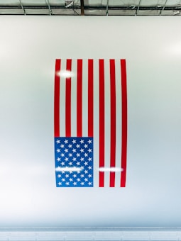 A stylized American flag with red vertical stripes and a blue square filled with white stars, placed on a white wall. The wall appears glossy, reflecting light from above. The flag is depicted in a vertical orientation instead of the traditional horizontal alignment.
